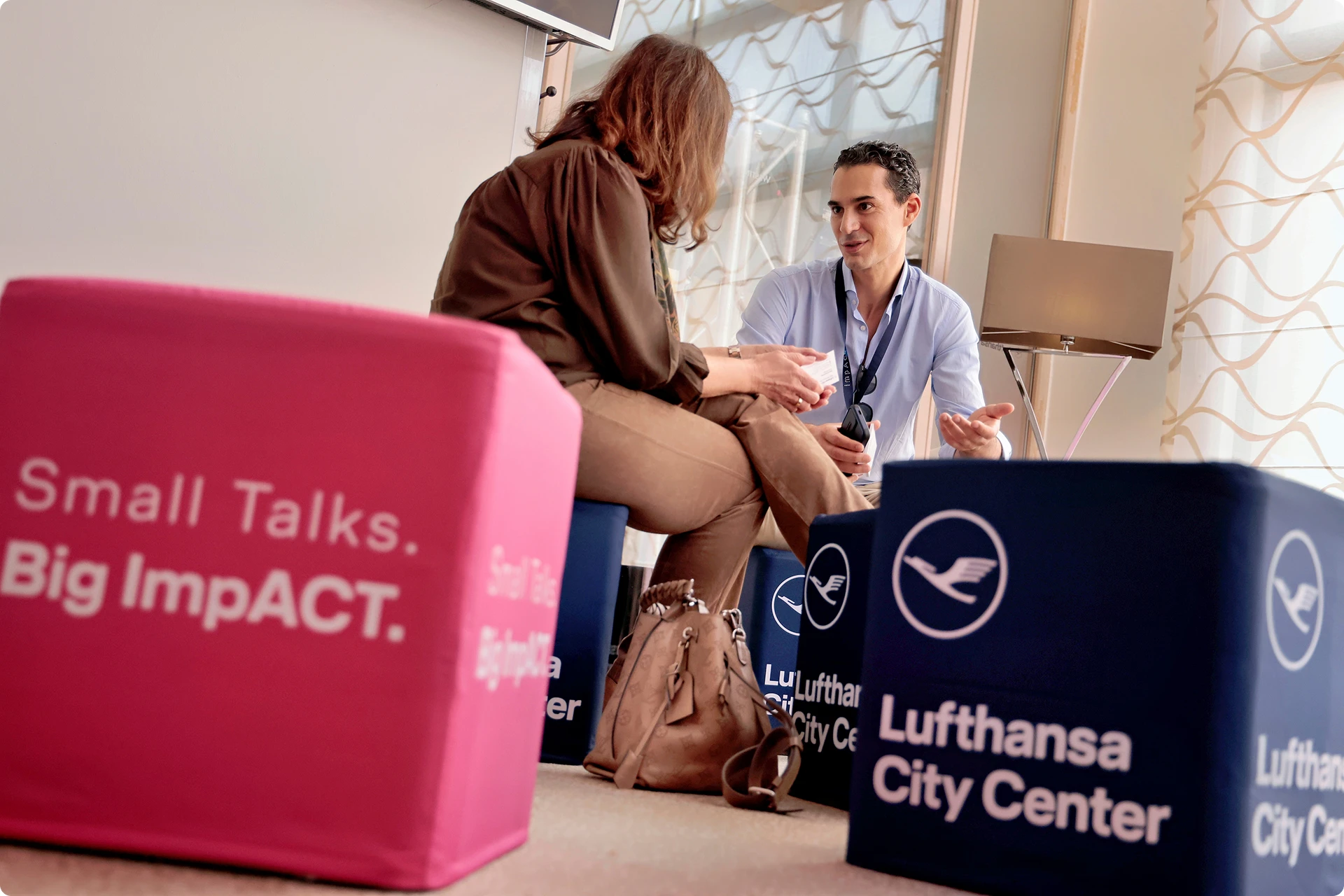 Two people seated and engaged in conversation in a lounge with cube seats branded with Lufthansa City Center and a pink cube with the text Small Talks. Big ImpACT.