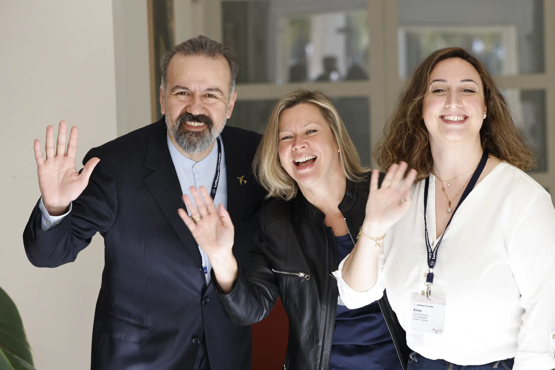 Three smiling people waving at the camera in an indoor setting.
