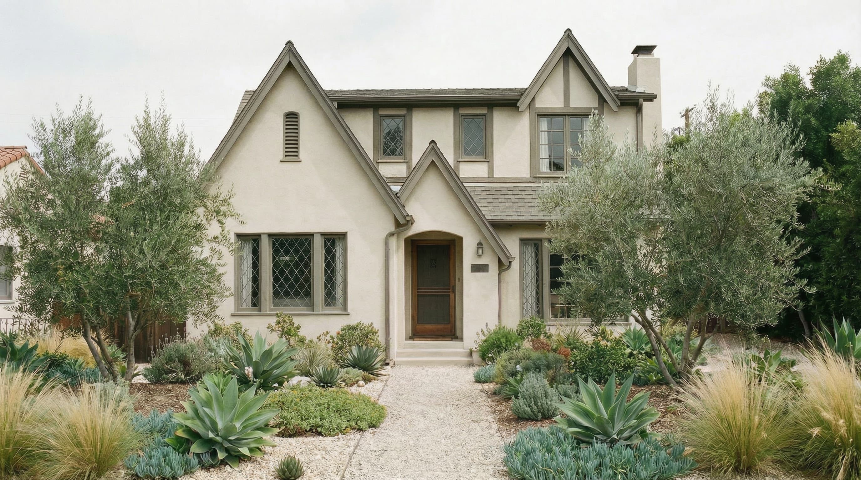 A beige two-story house with a steep roof, brown framed windows, and a wooden front door surrounded by a gravel path and lush drought-tolerant plants and shrubs.
