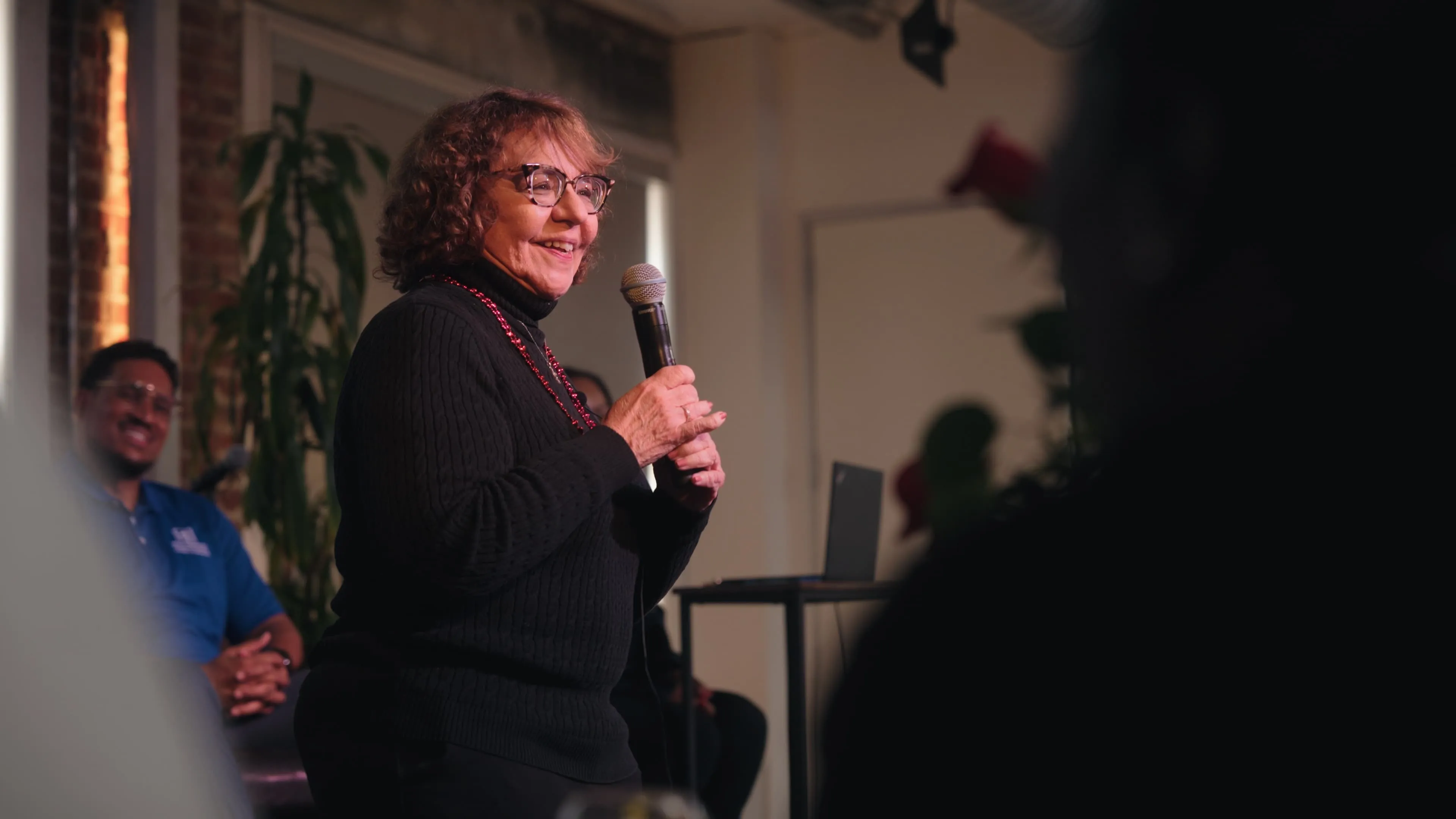 Older woman smiling and speaking into a microphone in a warmly lit indoor setting with a seated audience member in the background.