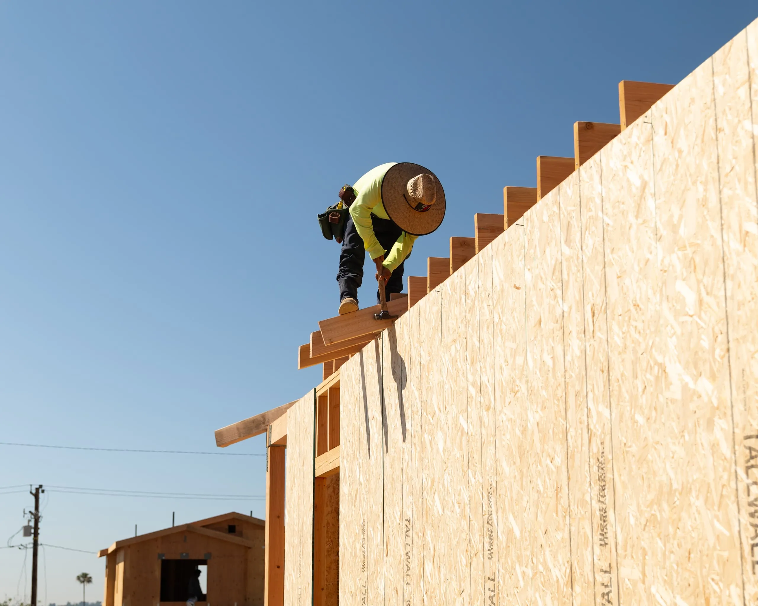 Construction worker wearing a straw hat and tool belt hammering wooden planks on roof frame under clear blue sky.