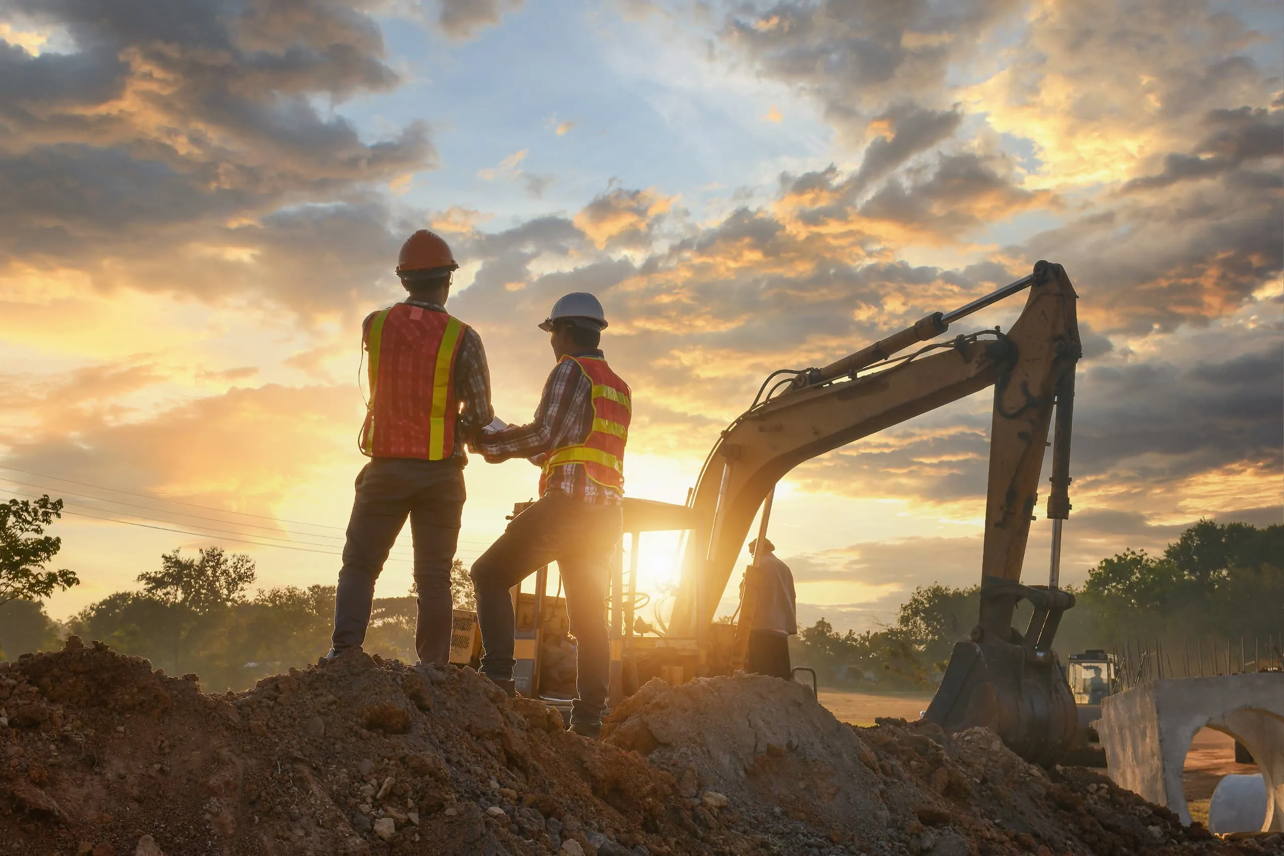 Two construction workers in safety vests and helmets standing on dirt near an excavator at sunset.