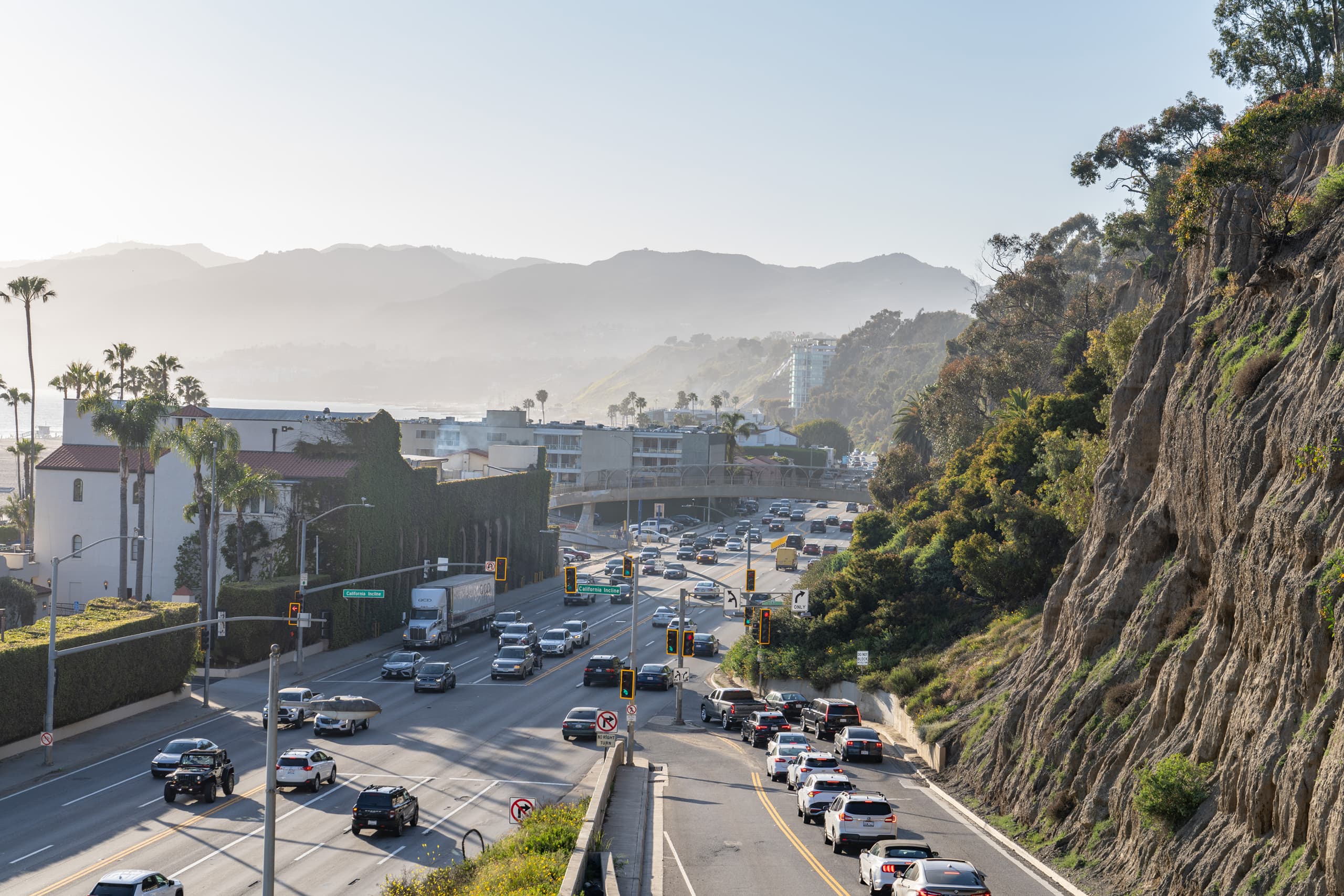 Busy multilane highway with cars and trucks near a rocky hillside and coastal buildings under a clear sky.