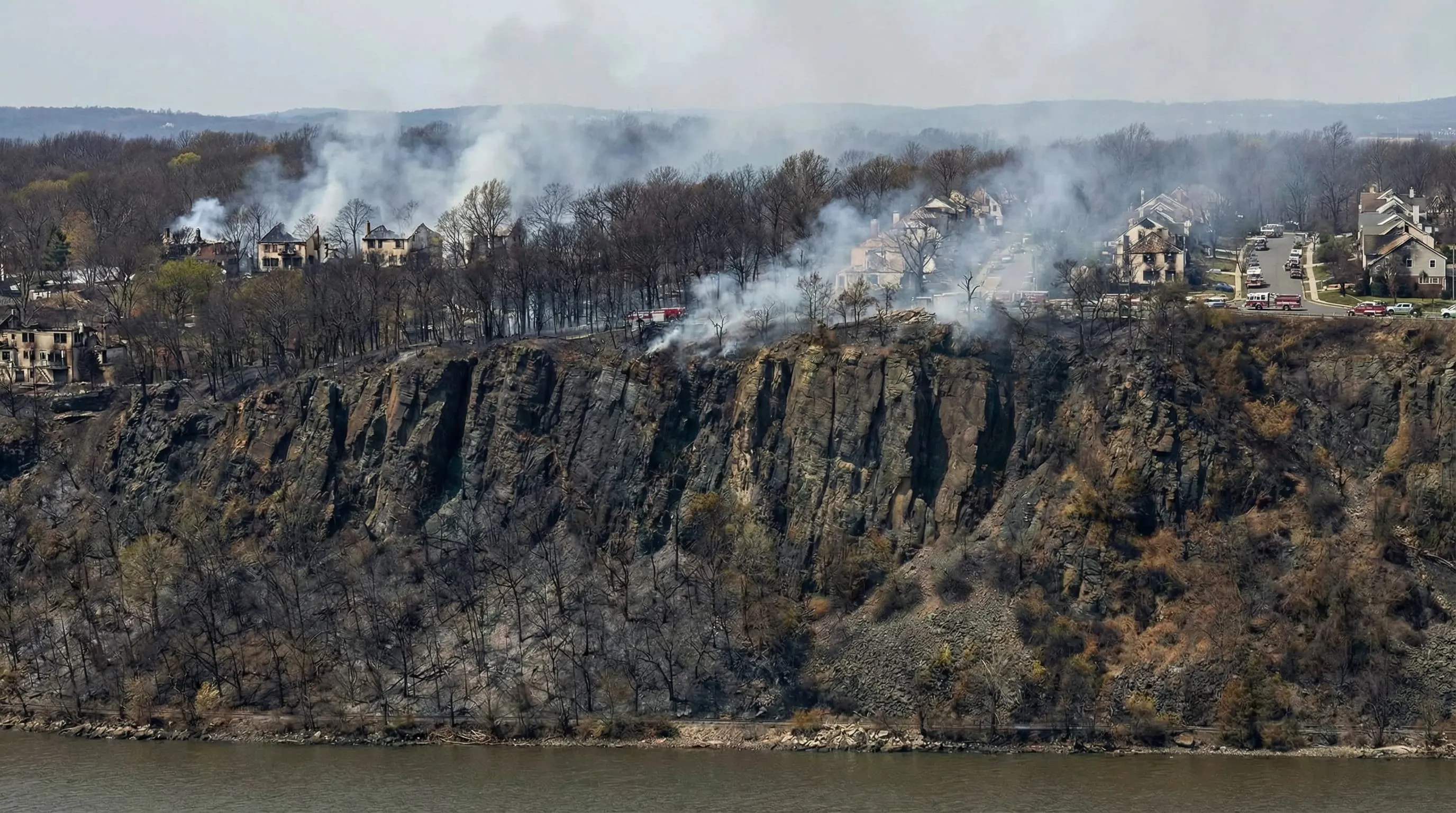 Cliffs overlooking a river with smoke rising from a fire burning in a wooded residential area at the top with fire trucks present.