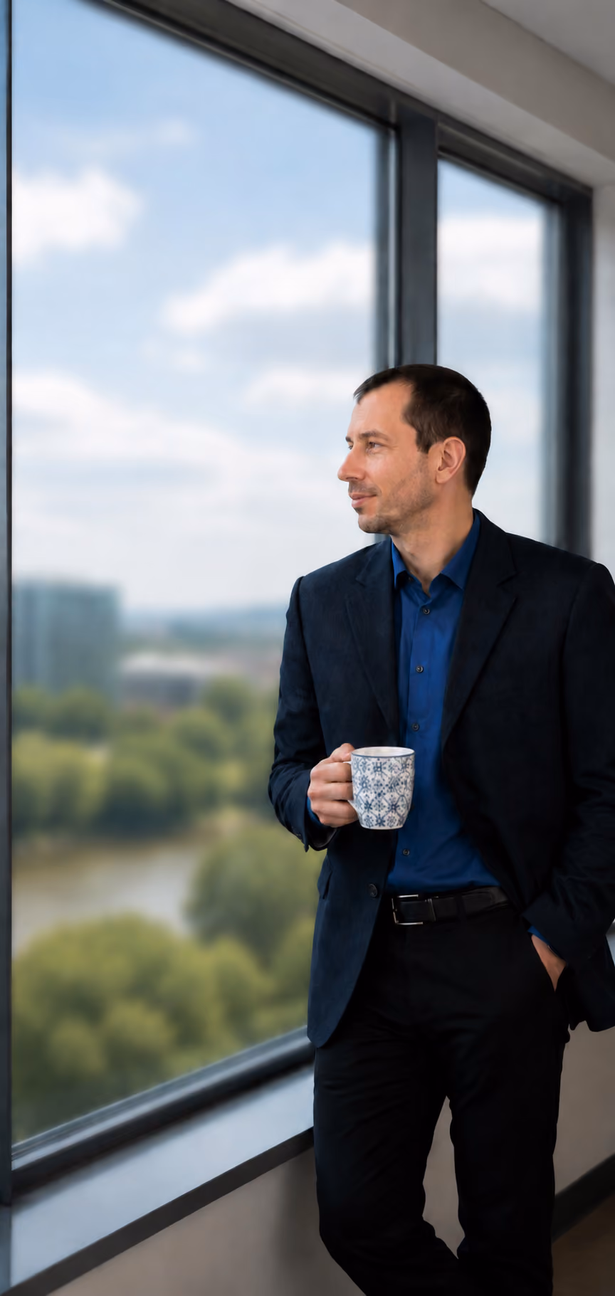 Ein Mann schaut lächelnd und entspannt aus einem Fenster und hält eine Tasse in der Hand.