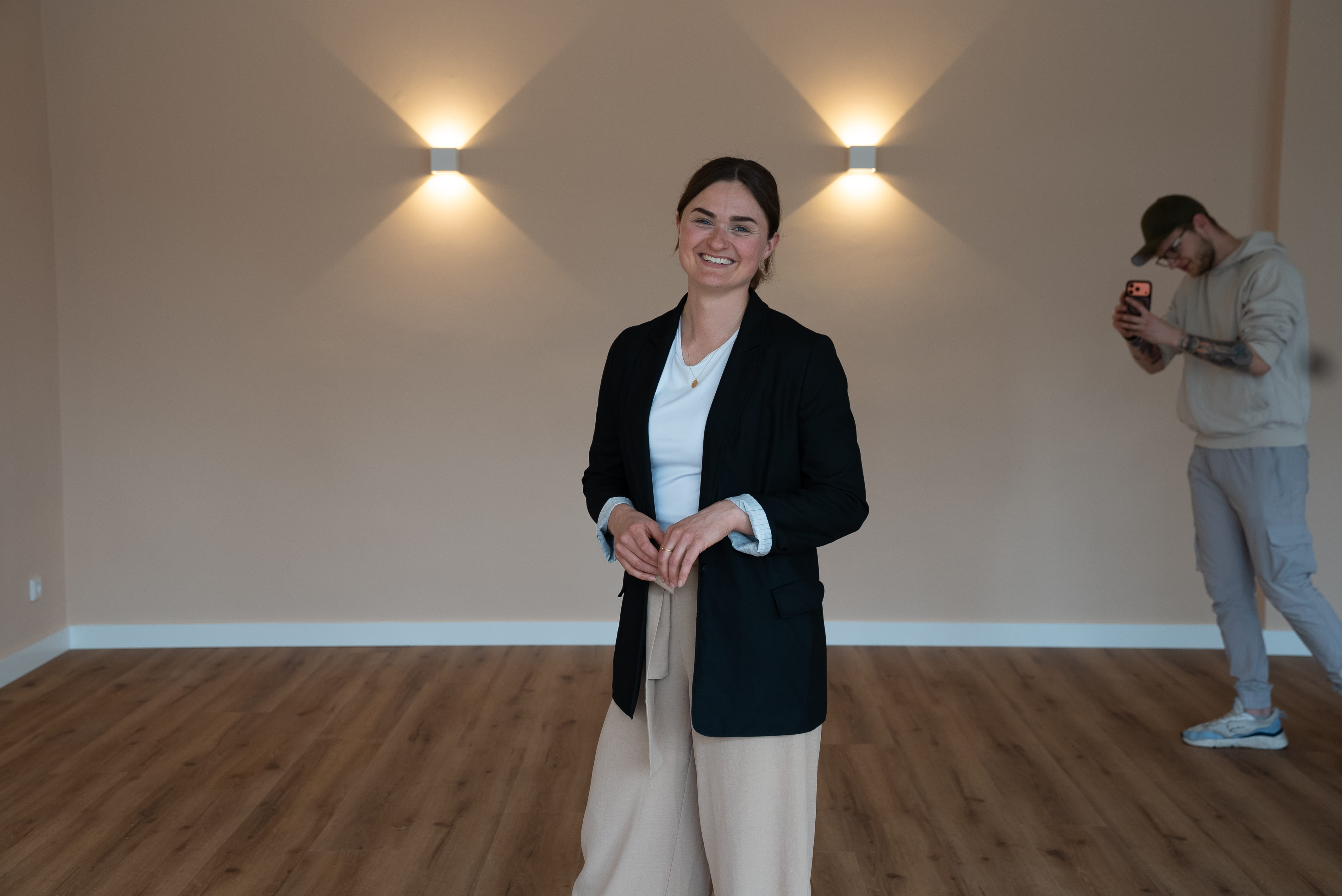 Lächelnde Frau in schwarzem Blazer und beigen Hosen steht in einem leeren Raum mit Holzfußboden, im Hintergrund fotografiert ein Mann mit Kapuzenpullover und Cap mit seinem Handy.
