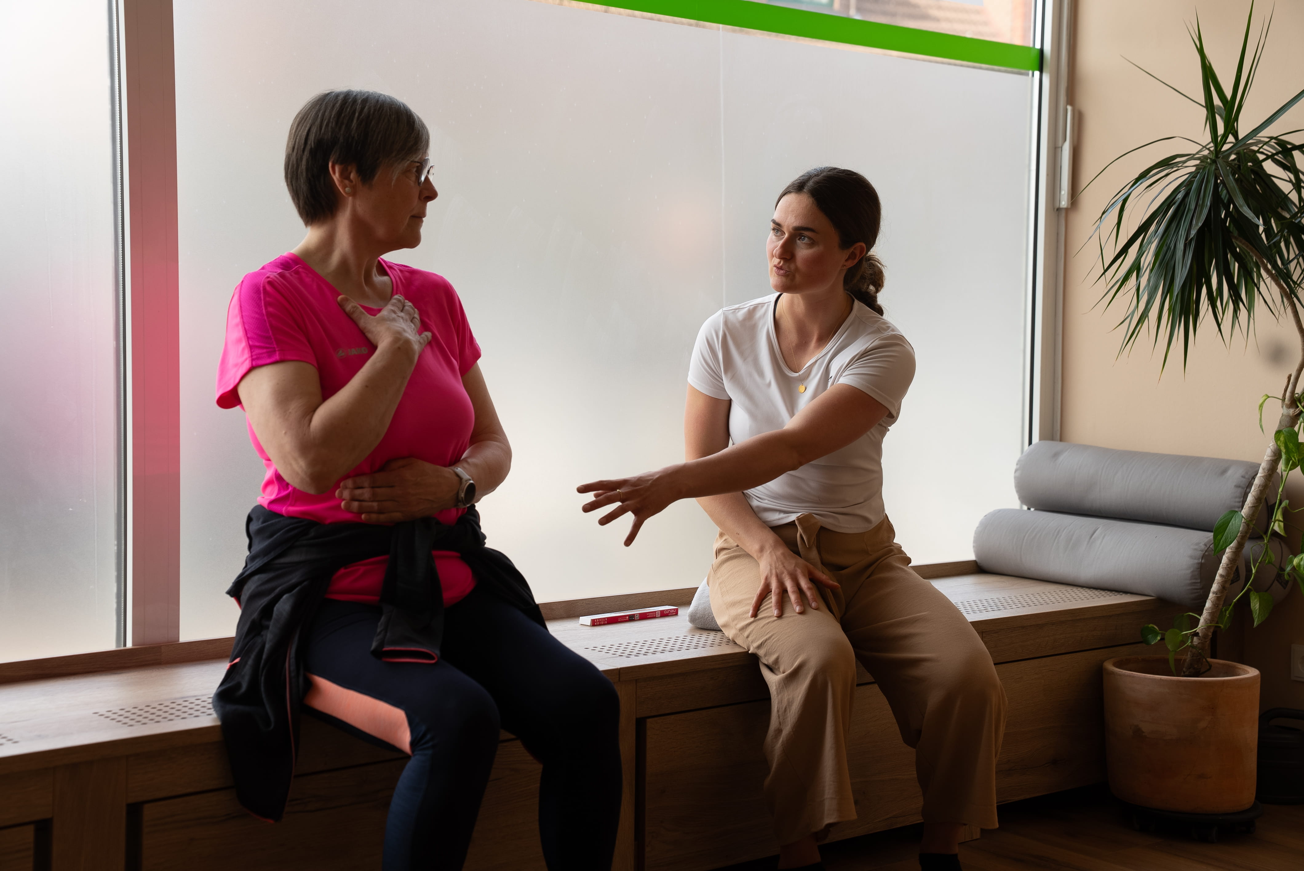 Zwei Frauen sitzen auf einer Holzbank, eine spricht und gestikuliert, die andere hält die Hand an Brust und Bauch.