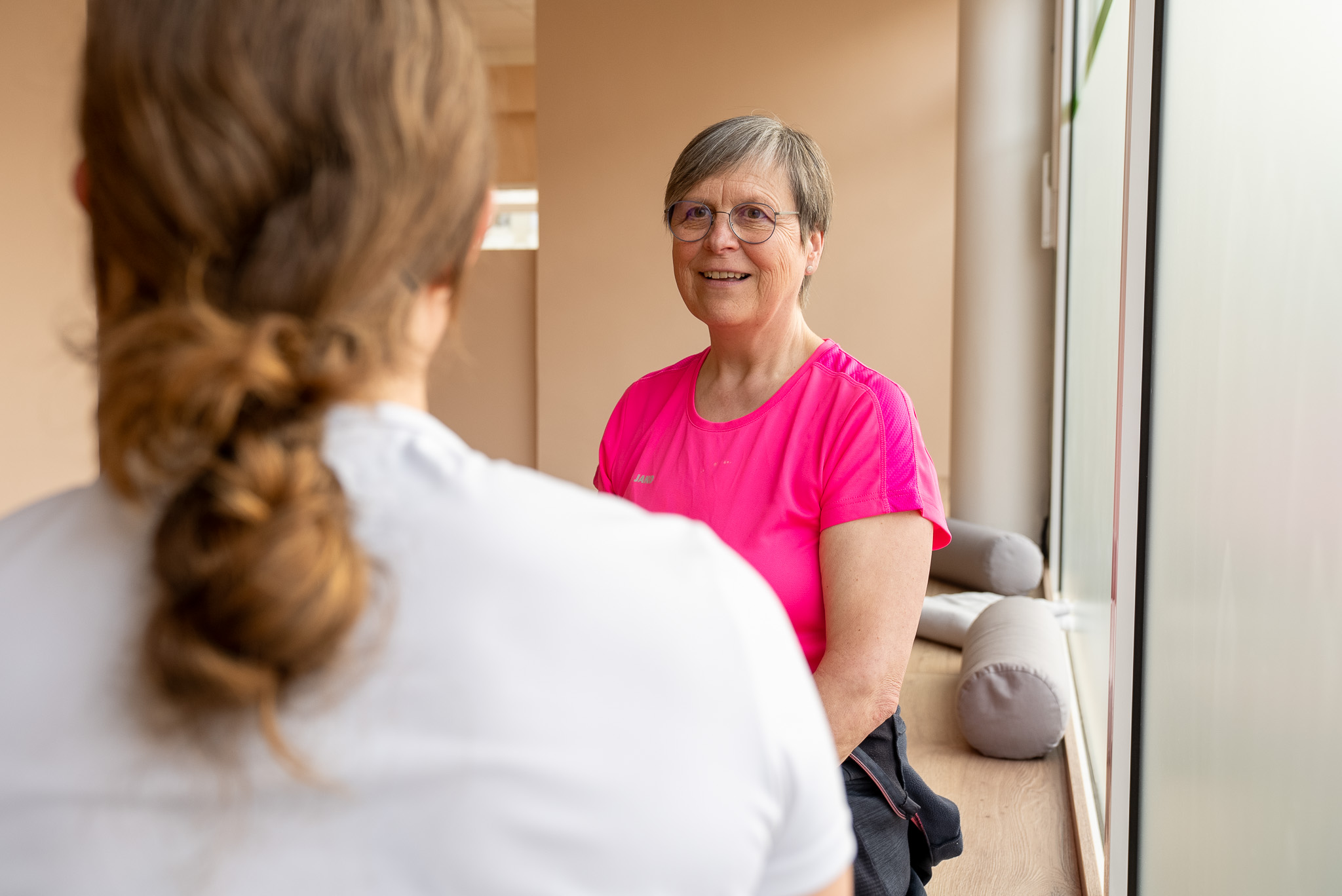 Eine lächelnde ältere Frau mit kurzen grauen Haaren und Brille, die in einem hellen Raum neben einem Fenster sitzt und eine pinke Sportshirt trägt, spricht mit einer Person mit Zopf.