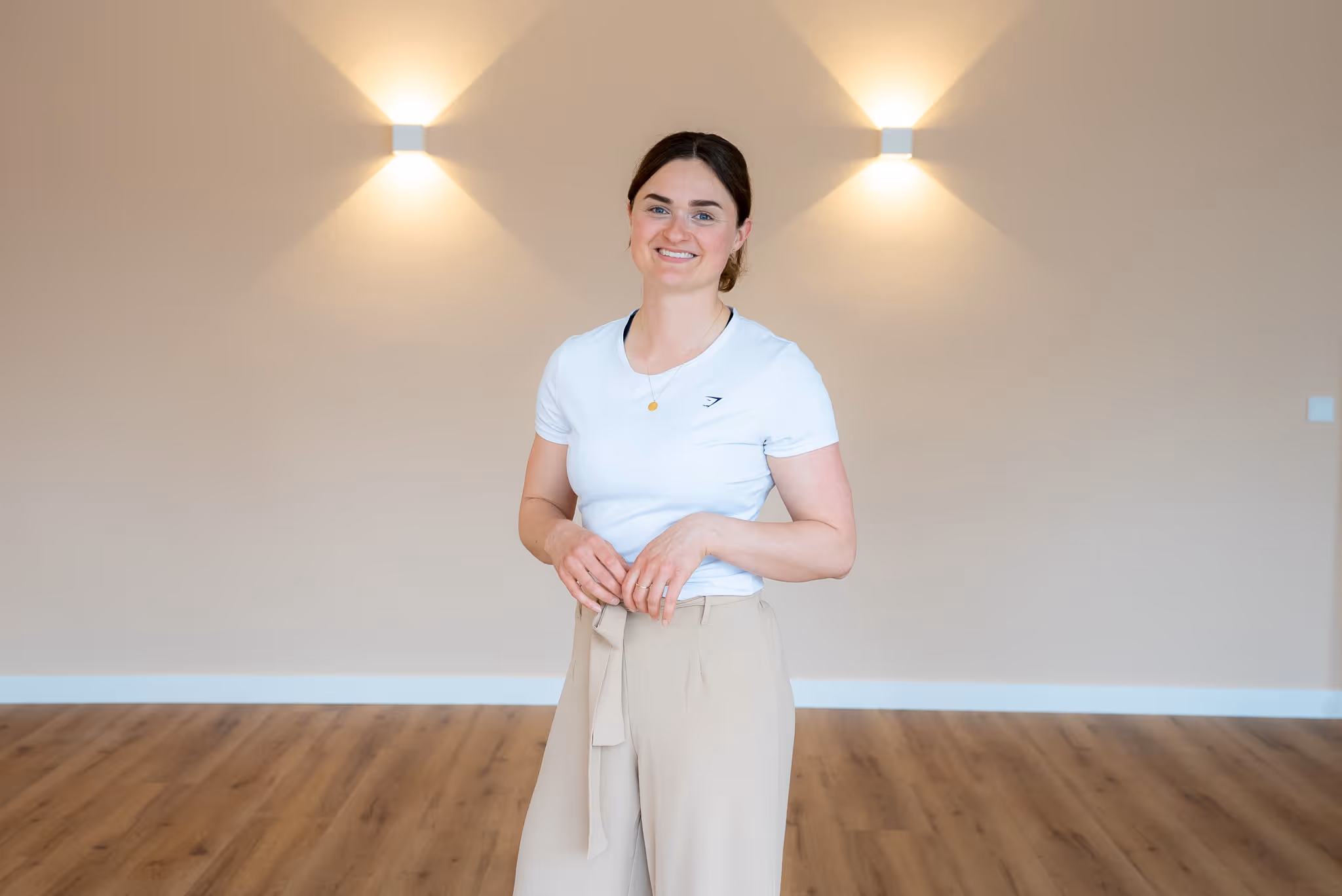 Junge Frau in weißem T-Shirt und beigen Hosen lächelt in einem minimalistischen Raum mit Holzboden und zwei Wandlampen.