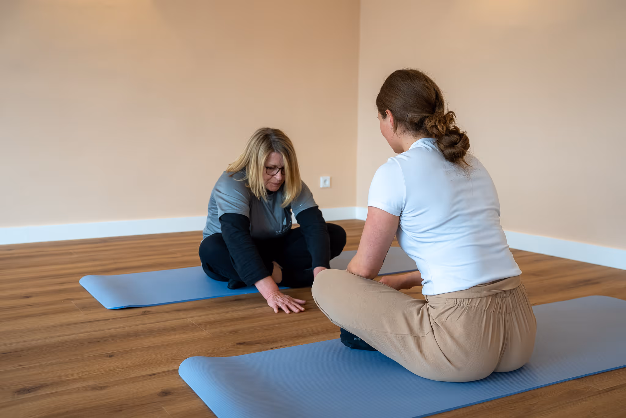 Zwei Frauen sitzen auf Yogamatten auf einem Holzboden, eine dehnt sich nach vorne, die andere schaut zu.