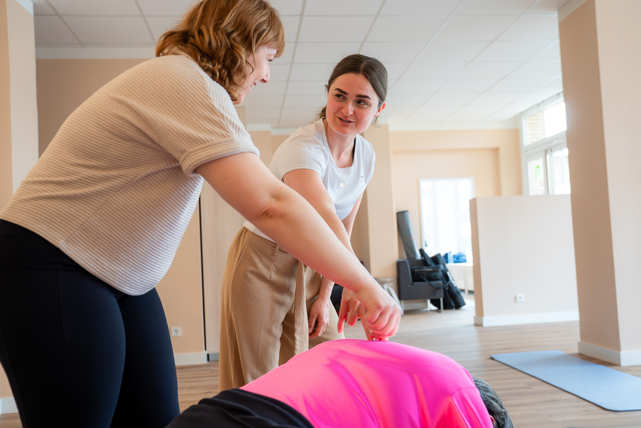 Zwei Frauen unterstützen eine dritte Frau bei einer Dehnübung in einem hellen Trainingsraum.