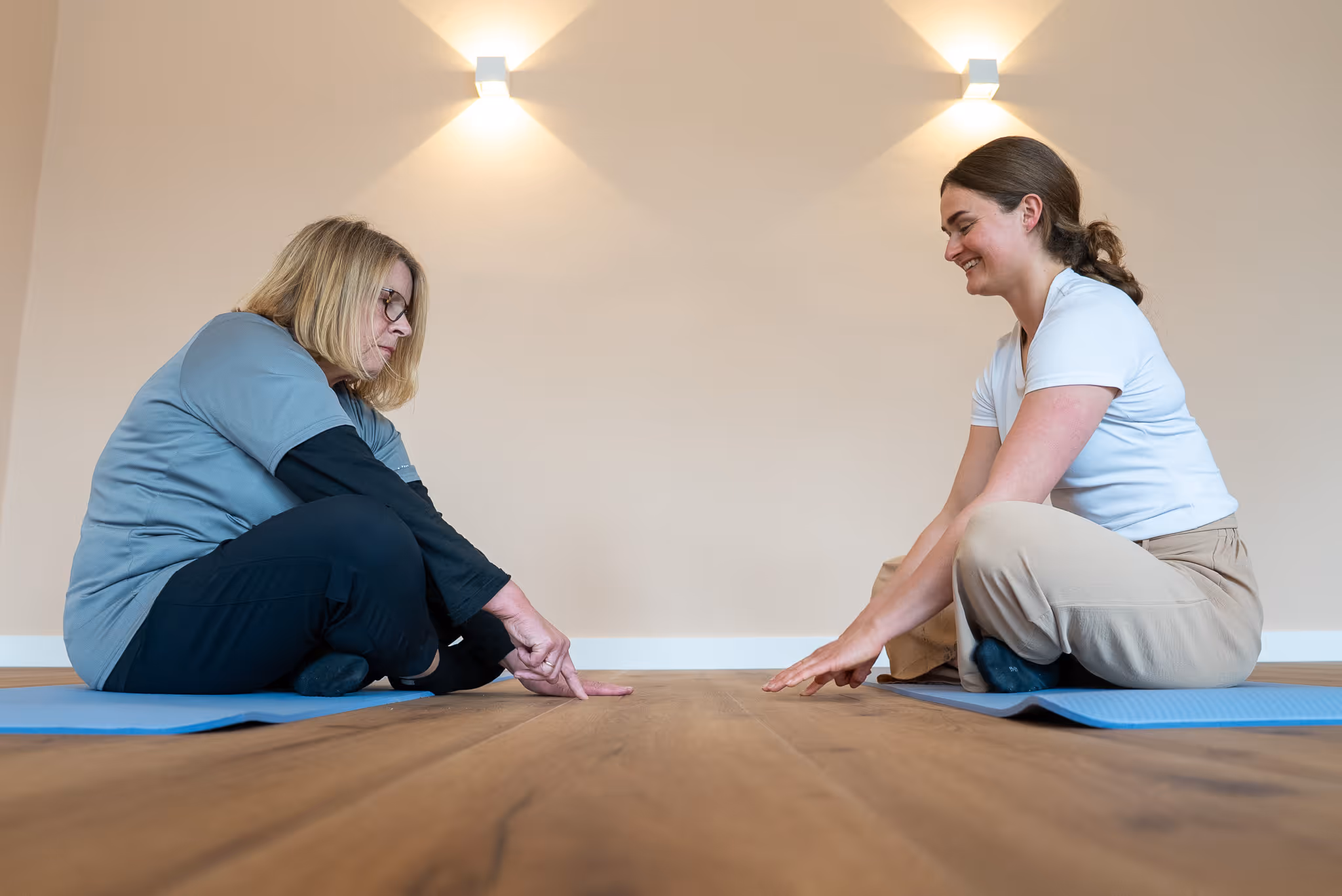Zwei Frauen sitzen auf Yogamatten im Schneidersitz und dehnen sich mit ausgestreckten Armen nach vorne.