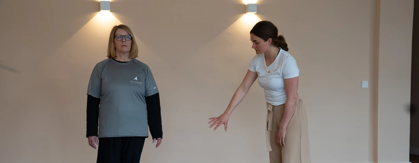 Zwei Frauen in einem hellen Raum, eine steht aufrecht mit grauem Shirt und Brille, die andere in weißem Shirt zeigt mit der Hand nach unten.