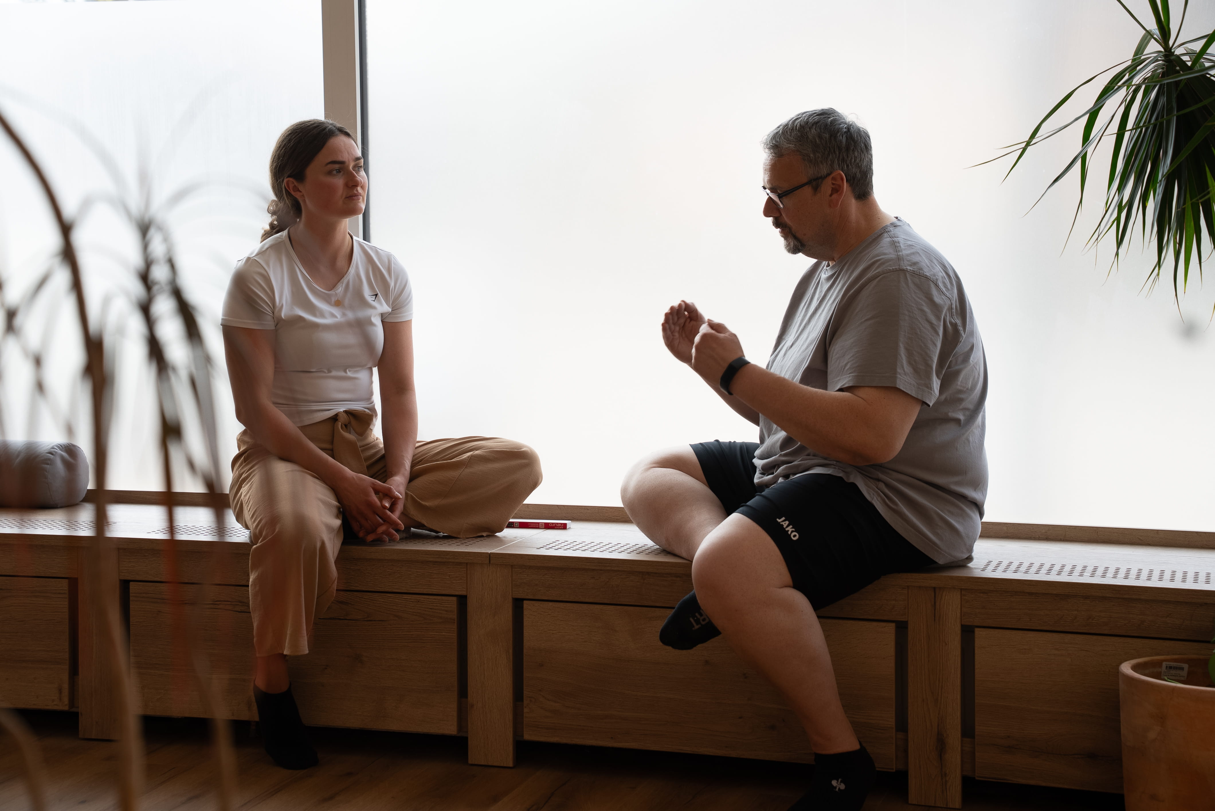 Eine Frau und ein Mann sitzen auf einer Holzbank vor einem großen Fenster und führen ein Gespräch.