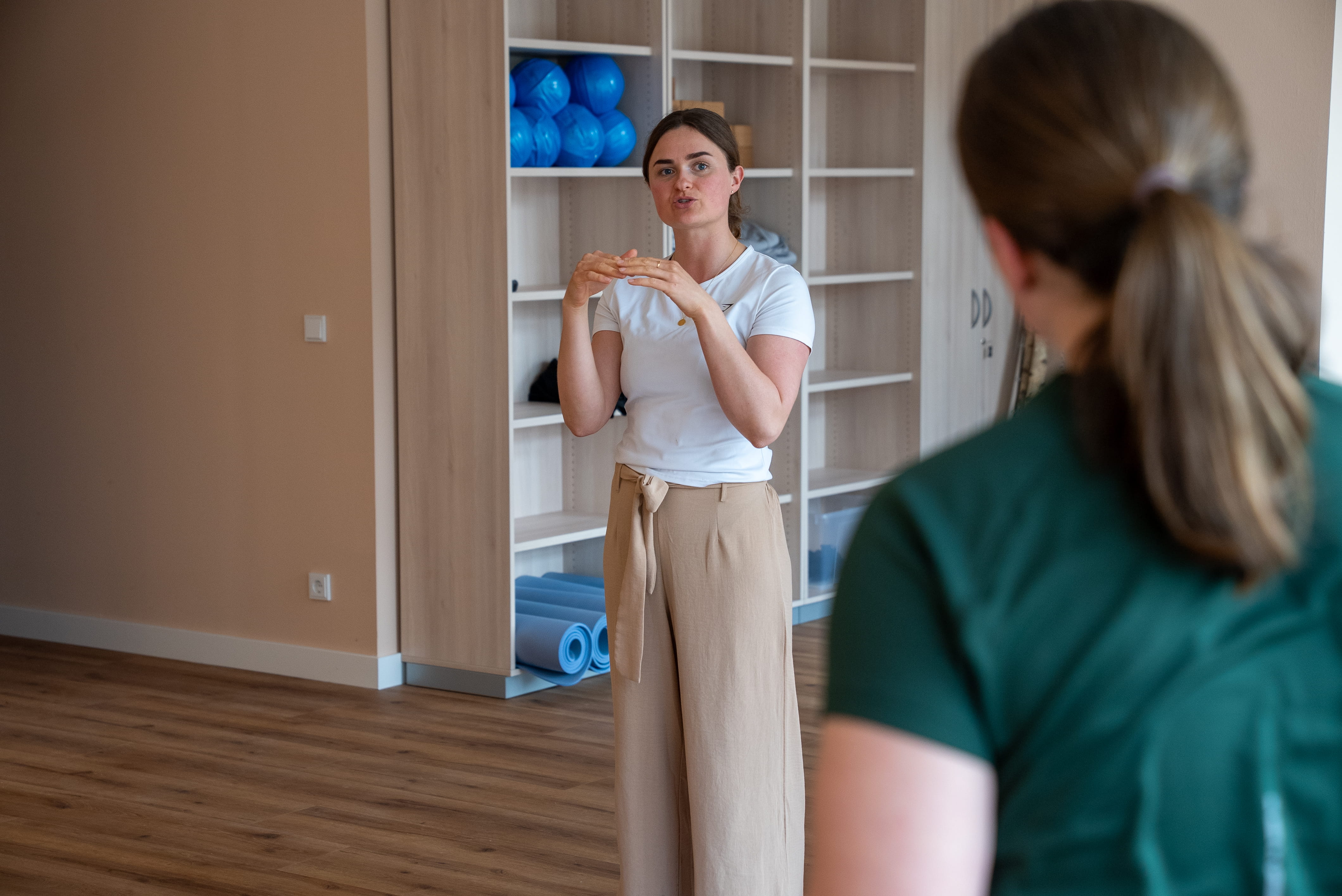 Frau in weißem T-Shirt und beiger Hose erklärt eine Sache, während eine andere Person ihr zuhört, im Hintergrund Regale mit blauen Helmen und Teppichen.