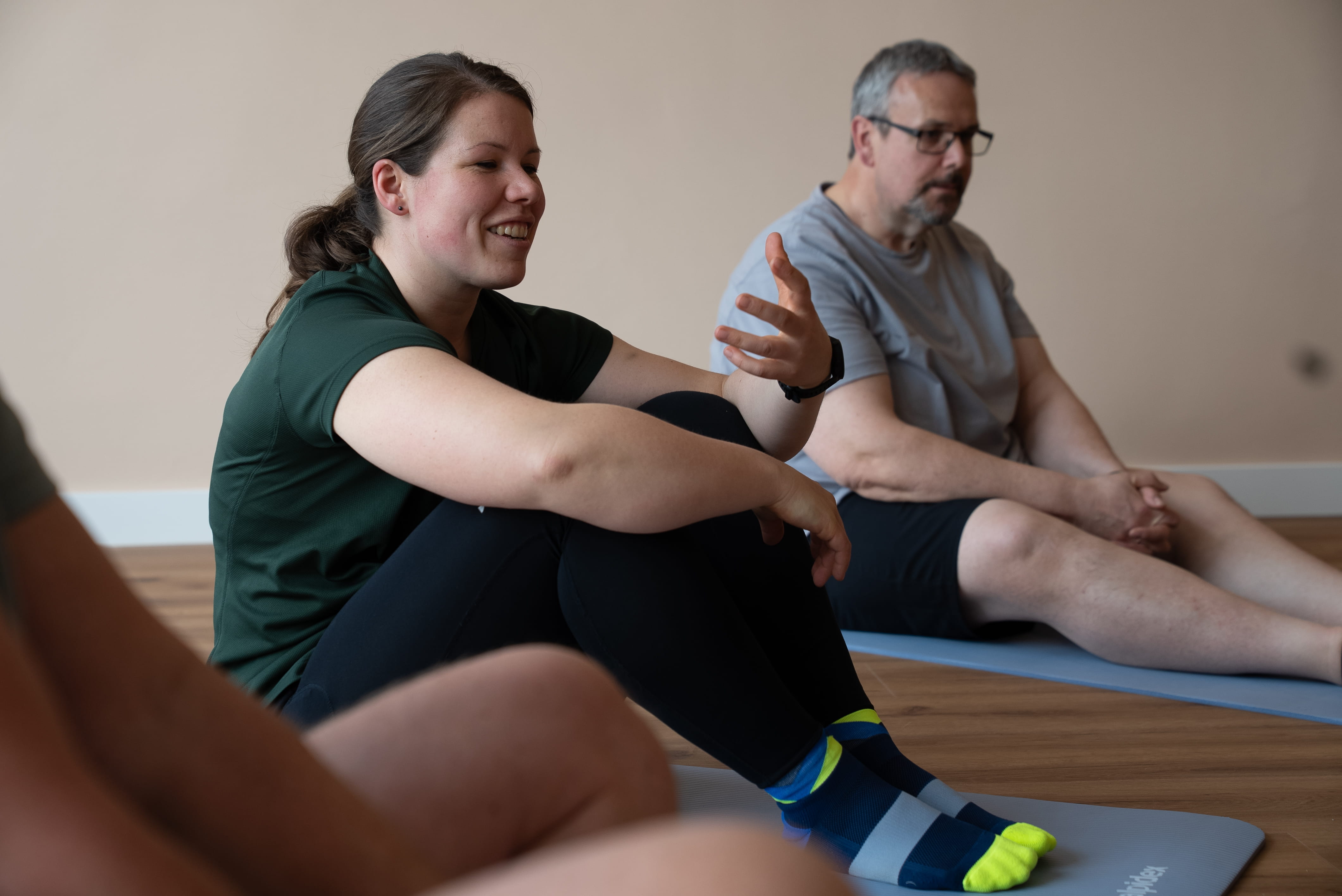 Frau und Mann sitzen auf Yogamatten auf dem Boden und unterhalten sich in einem hellen Raum.