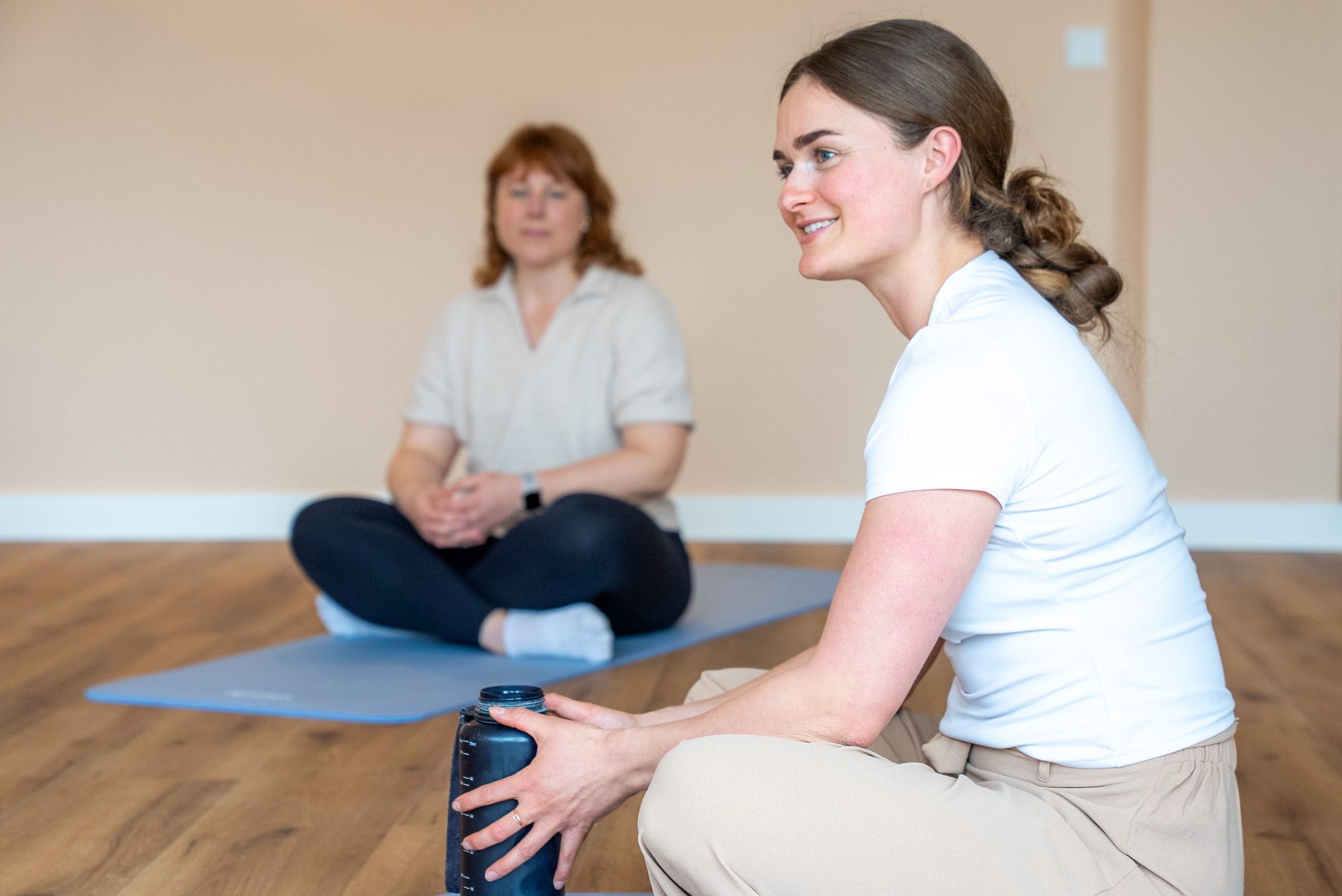 Zwei Frauen sitzen auf Yogamatten auf Holzboden, eine hält eine schwarze Trinkflasche und lächelt.