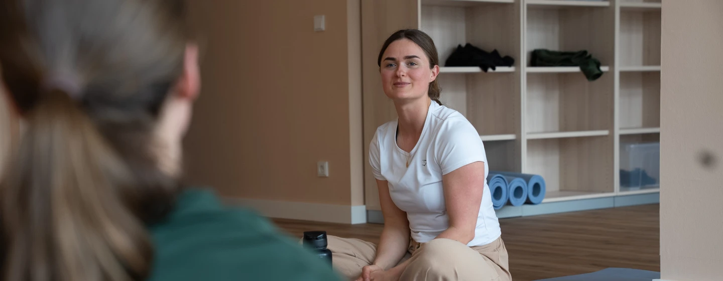 Frau sitzt auf Bodenmatte und unterhält sich in hellem Raum mit Regalen und Yogamatten.