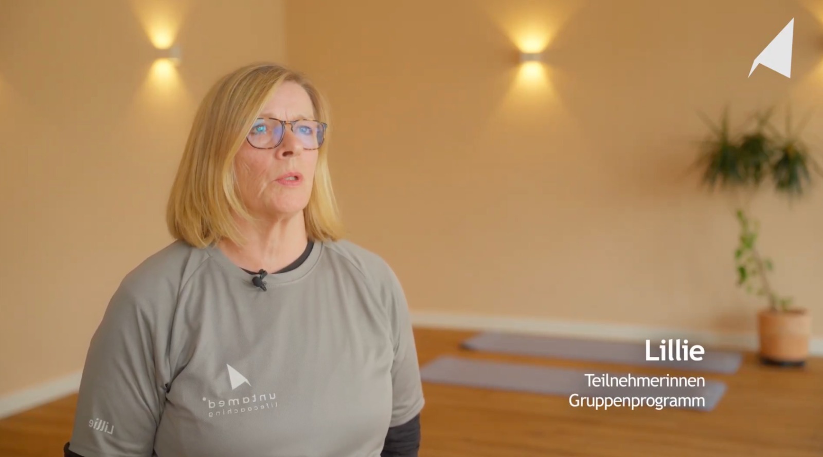 Frau mit Brille in grauem Shirt in einem Yogaraum mit Yogamatten und Pflanzen im Hintergrund.