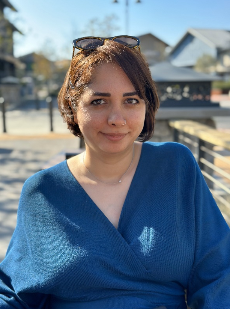 Woman with short brown hair, wearing sunglasses on her head and a blue V-neck top, smiling slightly outdoors on a sunny day.