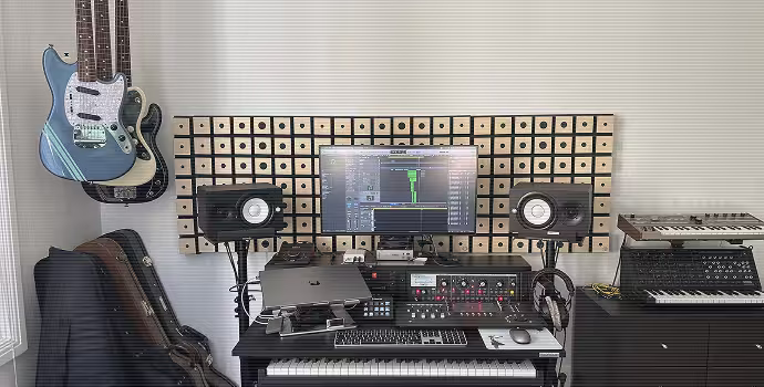 Configuration du studio de musique "La Crème Anglaise" avec deux guitares électriques montées sur le mur de gauche, un clavier, des haut-parleurs de contrôle audio, un écran d'ordinateur affichant un logiciel de musique, un équipement audio, des écouteurs et un synthétiseur à droite.