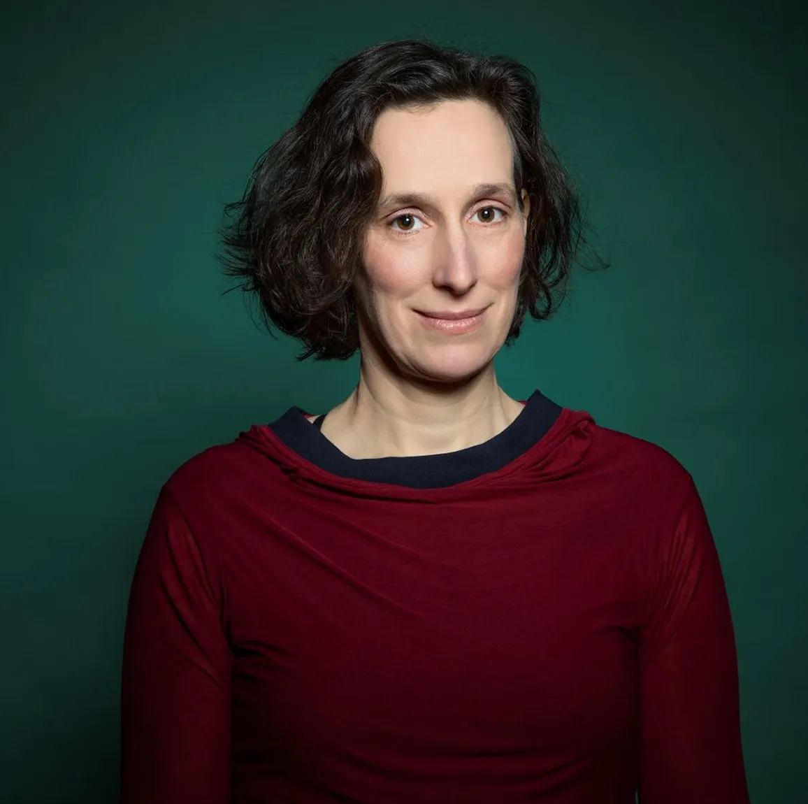 Frau mit kurzen, gewellten dunklen Haaren, die ein rotes Oberteil vor grünem Hintergrund trägt.