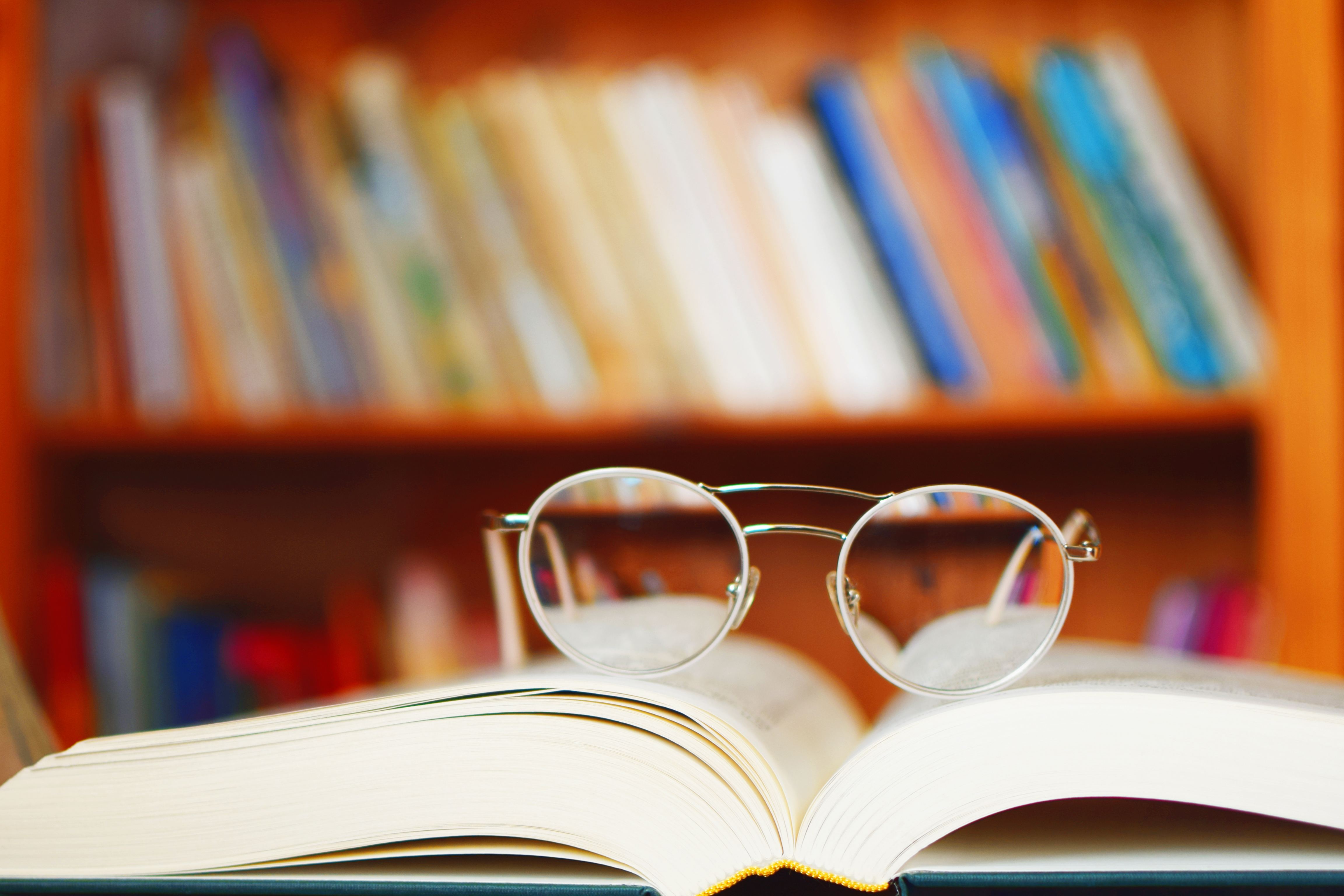 Runde Brille liegt auf einem geöffneten Buch, im Hintergrund unscharfes Bücherregal.