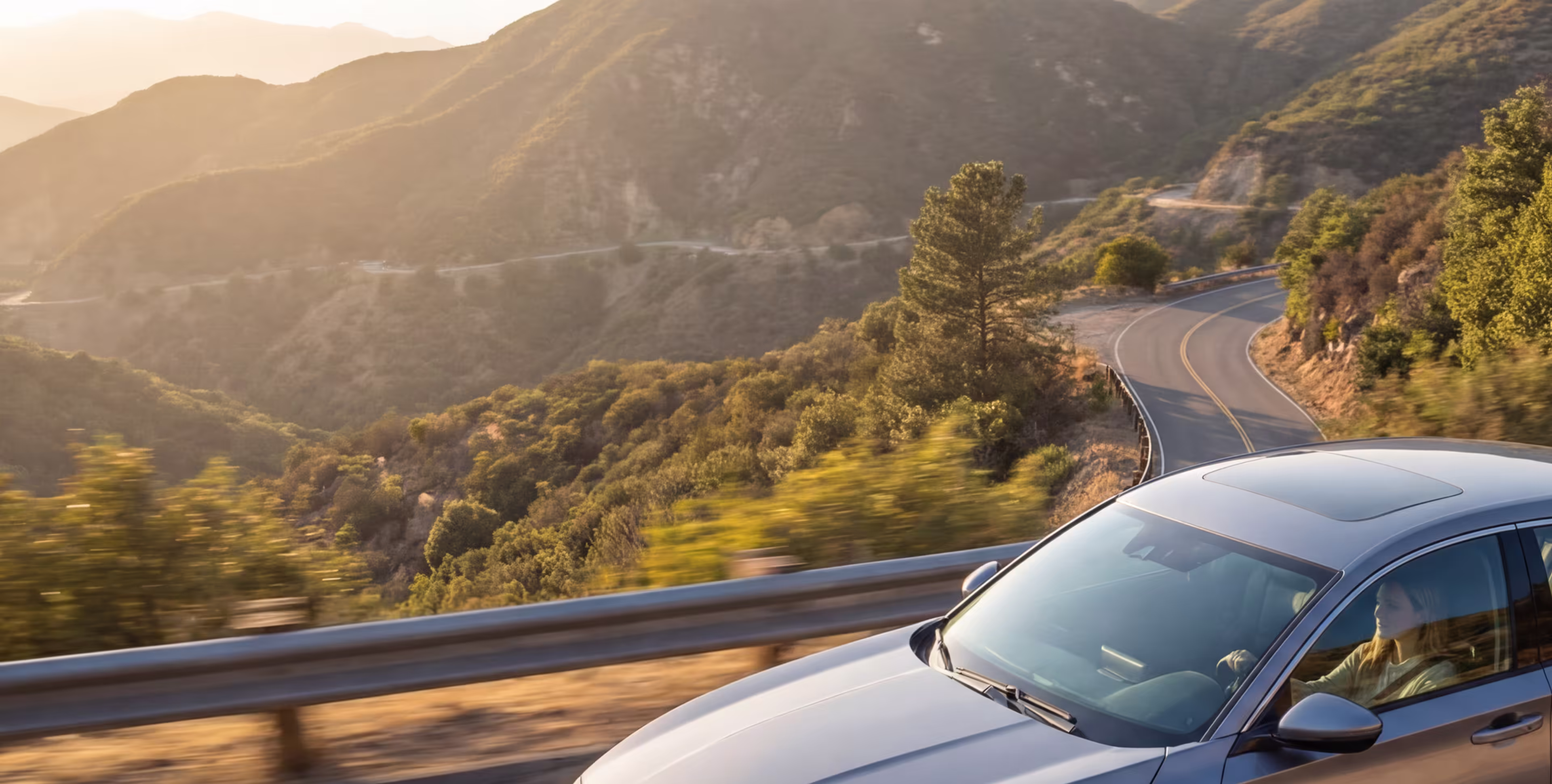 Silver car driving along a winding mountain road surrounded by sunlit trees and hills.