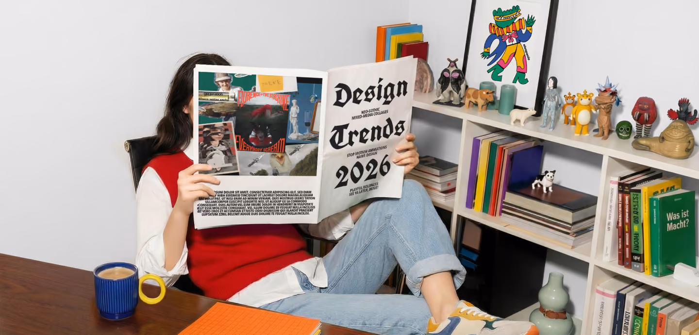 A person wearing a red vest and light jeans sitting at a desk, holding up a newspaper with the headline "Design Trends 2026" that covers their face.