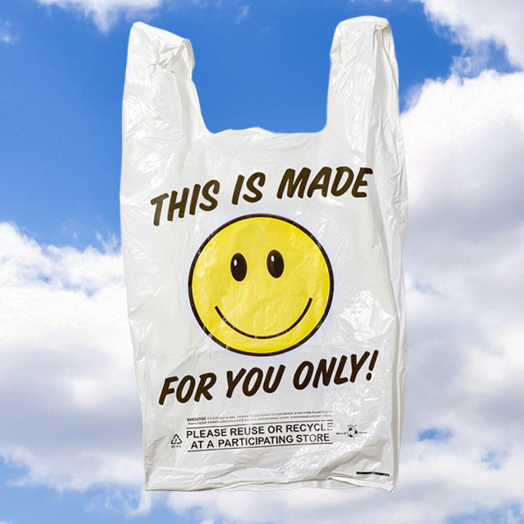 A white plastic grocery bag floating against a blue sky with fluffy clouds, printed with a yellow smiley face and the text "THIS IS MADE FOR YOU ONLY!".
