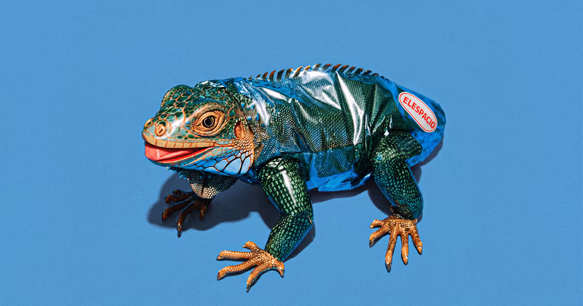 A green lizard wearing a metallic blue plastic suit featuring an "ELESPACIO" logo sticker, posed against a solid light blue background.