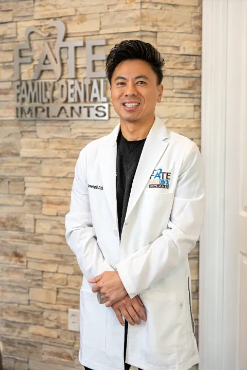 Smiling male dentist in a white coat standing in front of a stone wall with a 'Fate Family Dental Implants' sign.