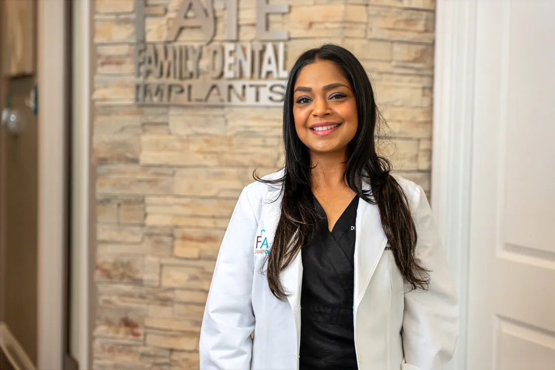 Smiling female dentist with long dark hair wearing a white coat standing in front of a stone wall with 'FAIE Family Dental Implants' sign.