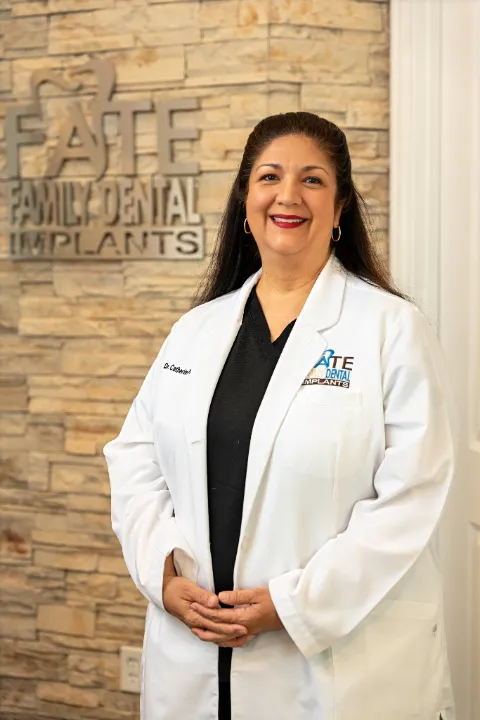 Female dentist wearing a white coat standing and smiling in front of a stone wall with FATE Family Dental Implants logo.