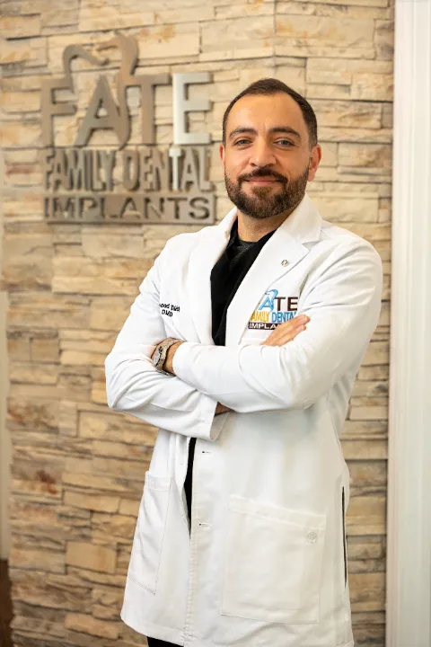 Male dentist in a white coat standing with arms crossed in front of a brick wall with the sign 'FATE Family Dental Implants'.
