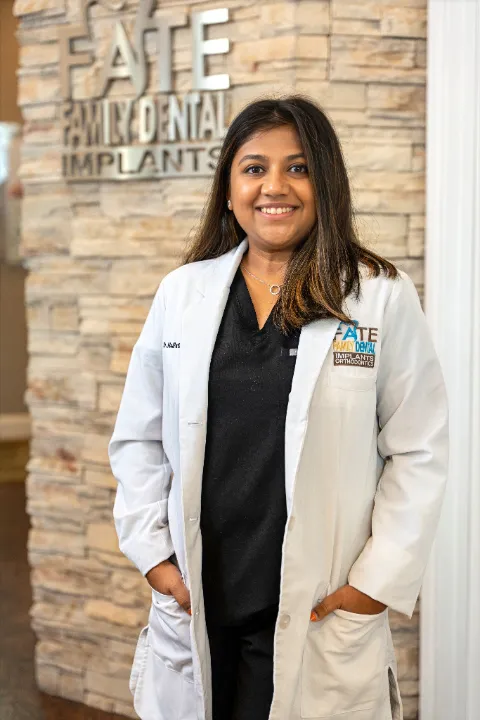 Smiling female dentist wearing a white coat with 'Fate Family Dental Implants Orthodontics' logo standing in front of a stone wall with the clinic's sign.