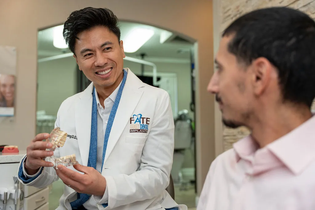 Smiling dentist in a white coat showing a dental implant model to a male patient in a dental office.