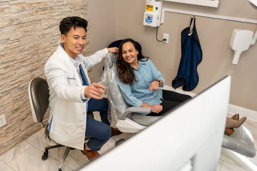 Dentist in a white coat pointing at a screen while a female patient sits in a dental chair smiling.