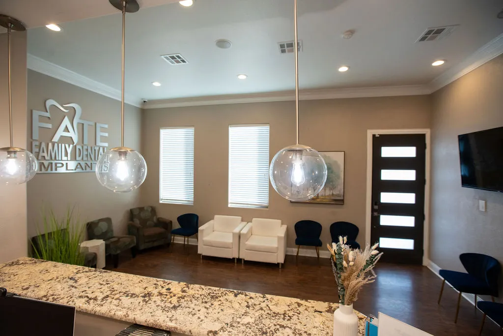 Modern dental office waiting room with white and dark blue chairs, a granite reception counter, hanging globe lights, and a wall-mounted TV.