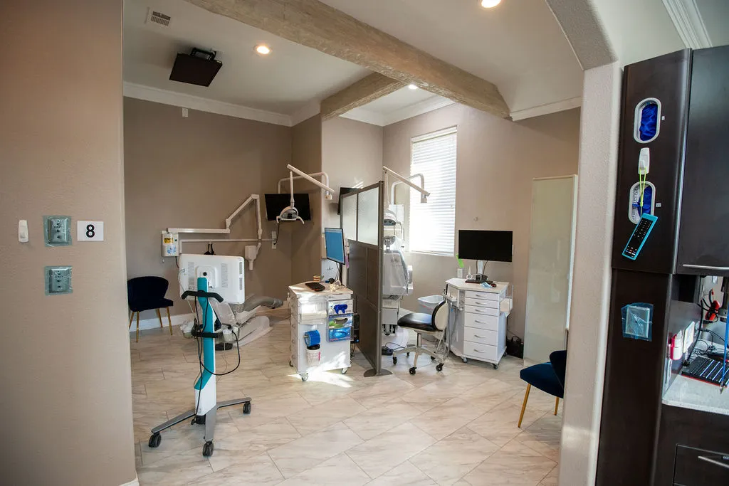 Modern dental office with two dental chairs, computer monitors, equipment carts, and beige walls under natural light from a window.
