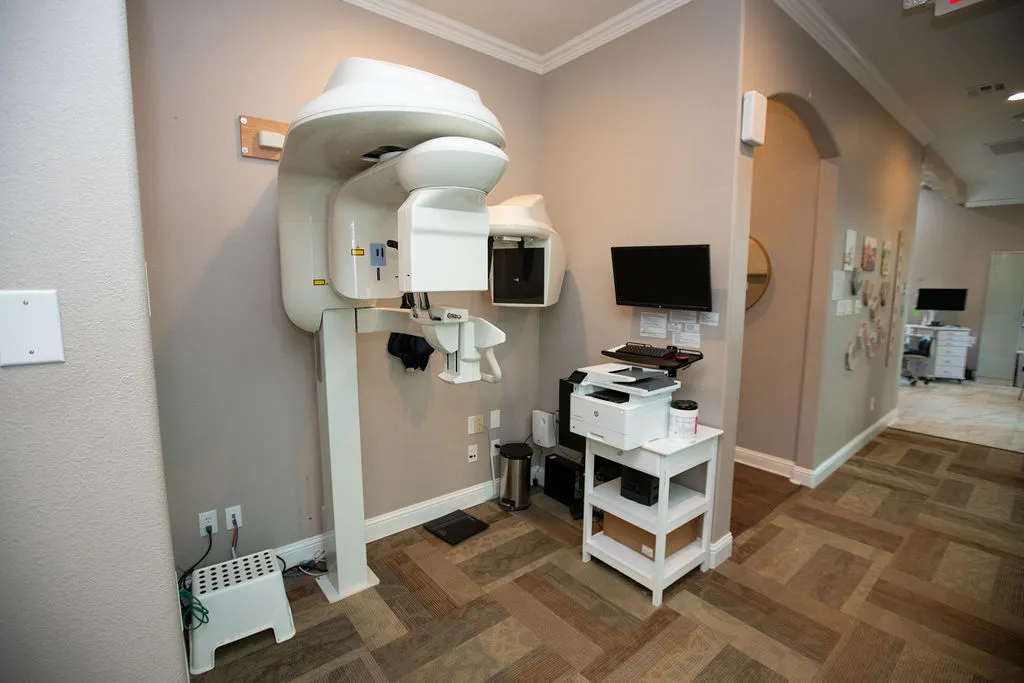 Dental or medical X-ray machine mounted on a wall in a clean, modern office with a computer monitor and printer on a small white table nearby.