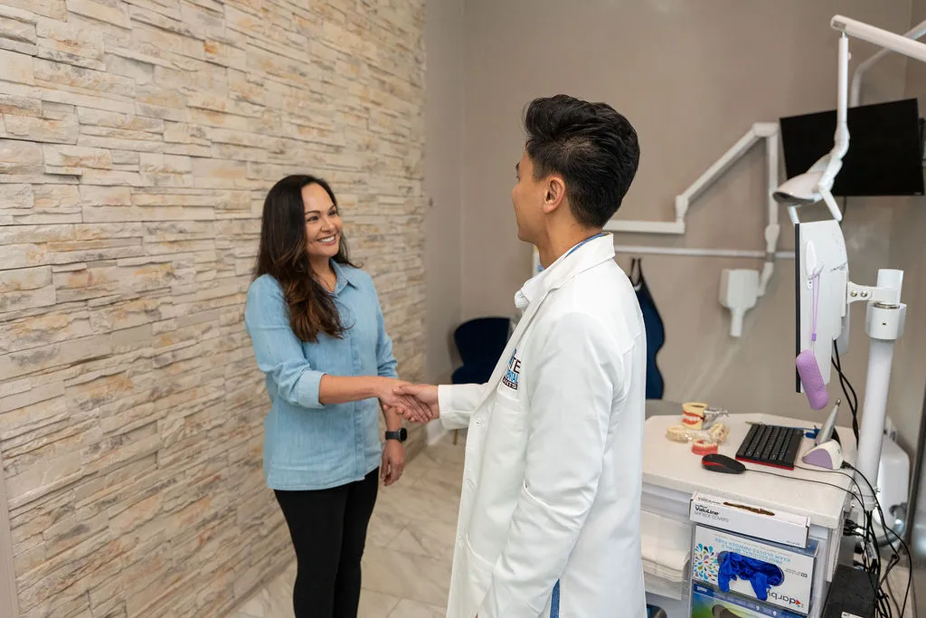 A female patient and a male doctor shaking hands in a medical office with dental models and computer equipment.