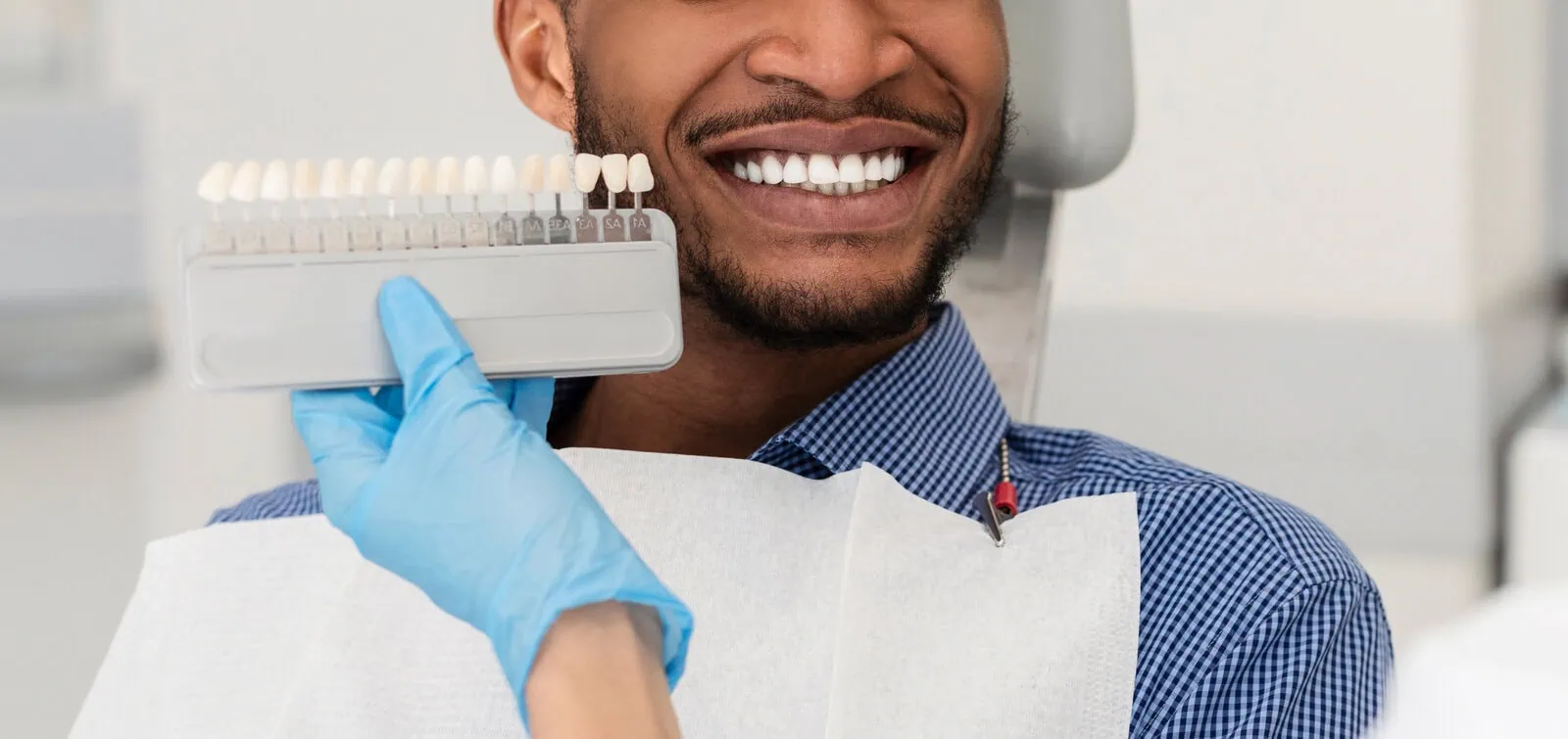Man in dental chair smiling while a gloved hand holds a tooth shade guide next to his teeth for color matching.