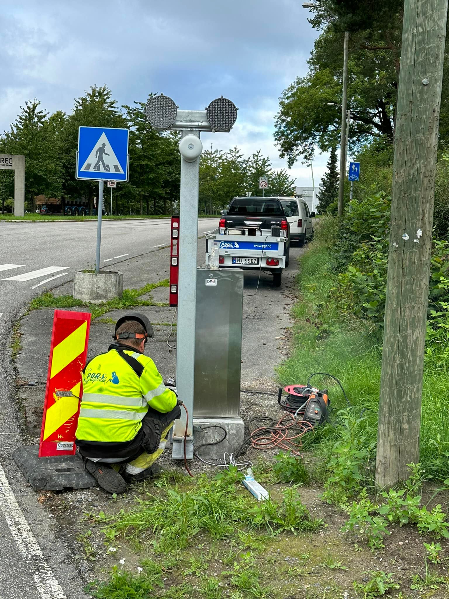 Worker in a yellow and black safety jacket kneeling on the ground next to a metal post with circular lights by the roadside under a pedestrian crossing sign.