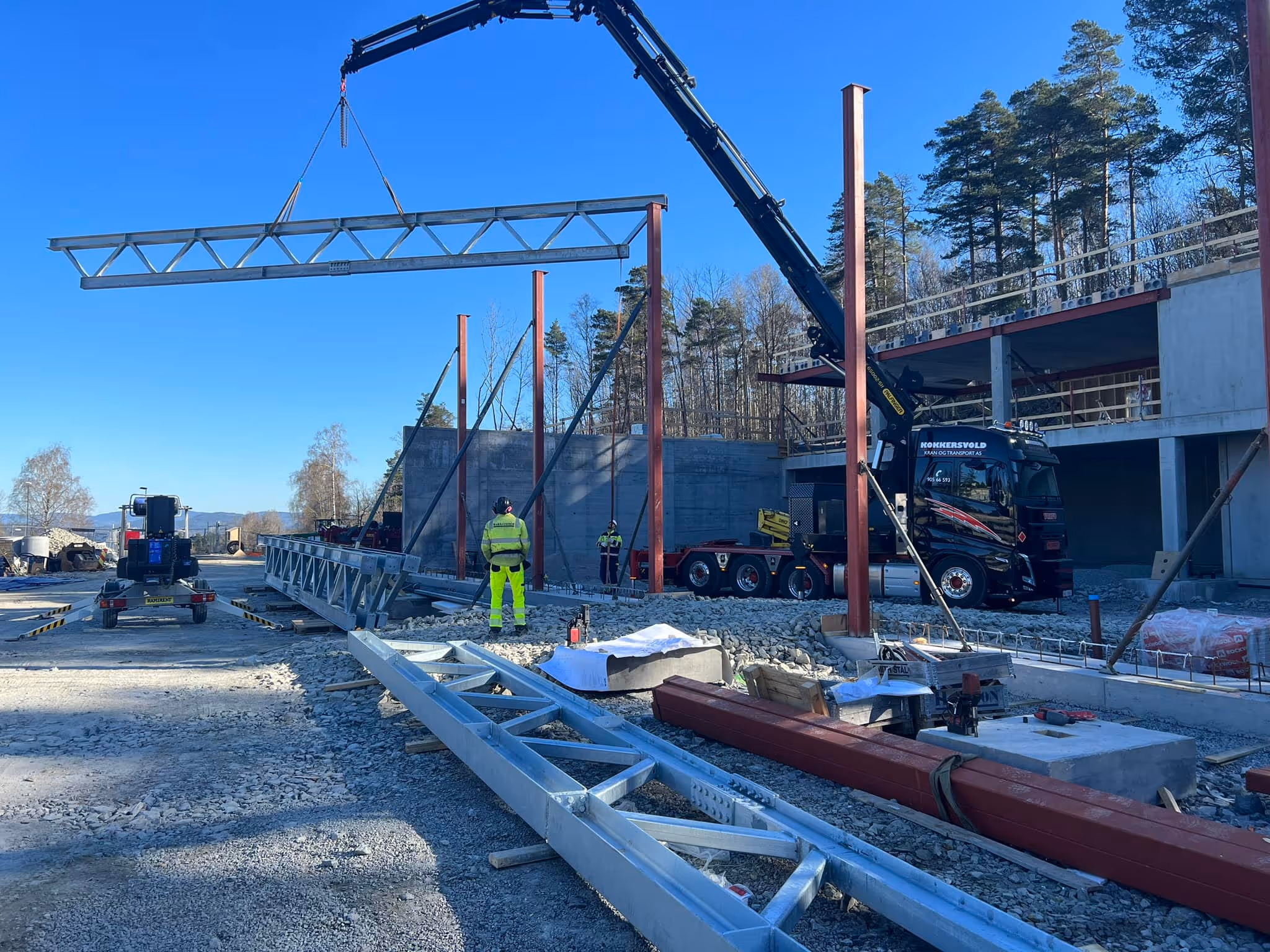 Construction site with a crane lifting a large metal truss while workers in high-visibility gear guide it near a partially built structure.