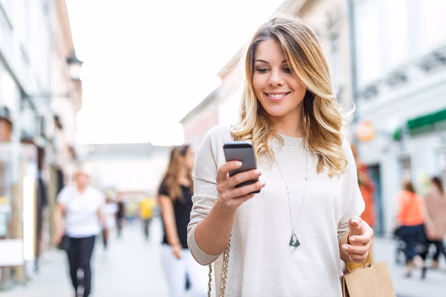 Smiling woman in white sweater looking at smartphone while walking down a busy city street.