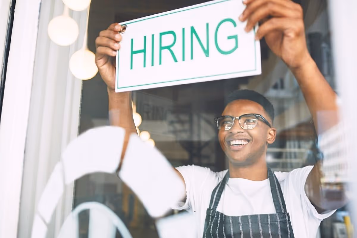 Smiling man in glasses and apron hanging a hiring sign on a storefront window.