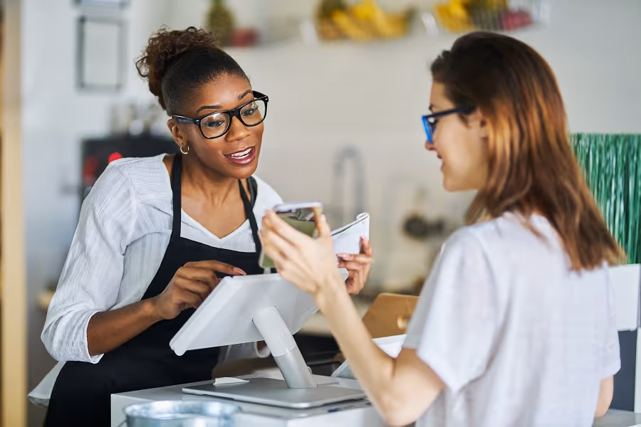 Smiling cashier in apron helping customer holding phone at a checkout counter.