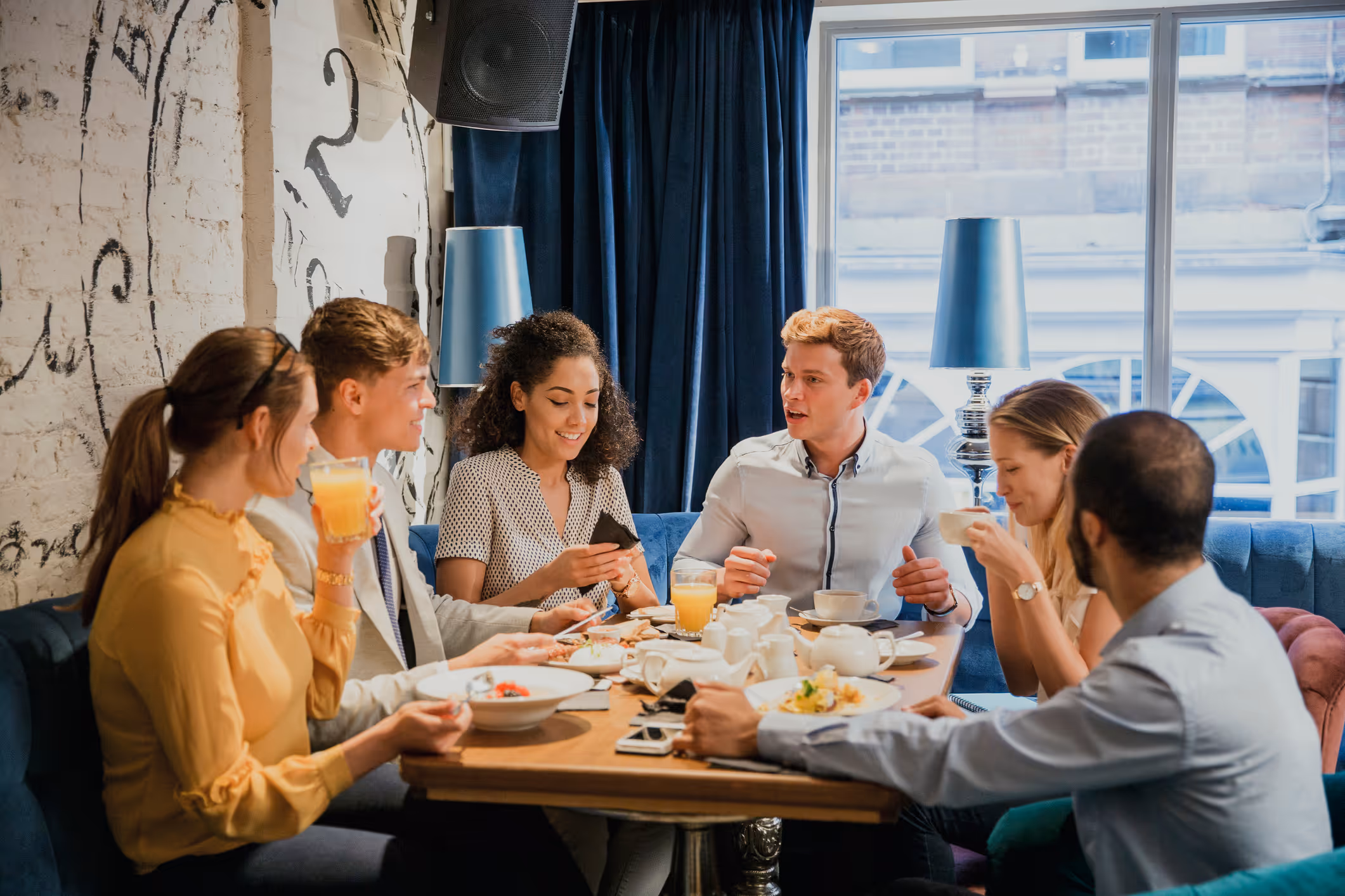 Group of six young adults enjoying food and drinks while seated at a restaurant table near a window.