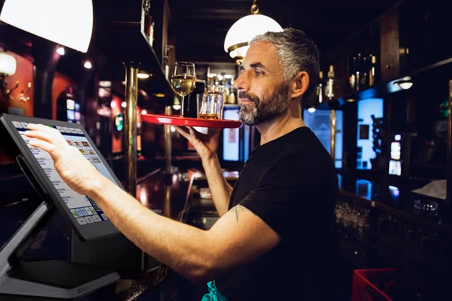 Restaurant owner carrying drinks on a tray while using a touchscreen POS system at the bar.